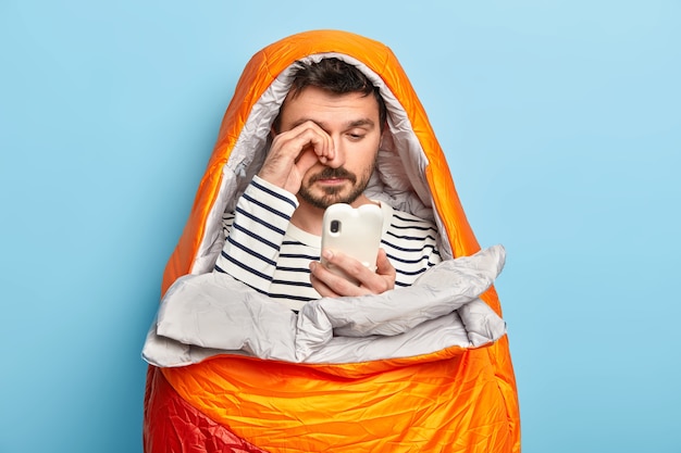 Free photo tired male camper rubs eyes, uses mobile phone, tries to connect to intenrnet in wild nature, poses in sleeping bag, has all necessary equipment needed for camping