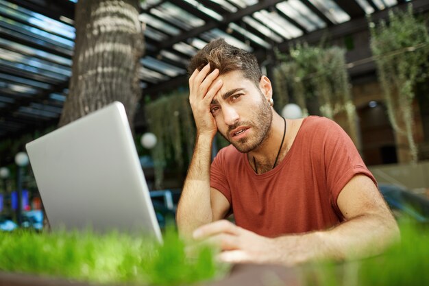 Tired male programmer, IT guy looking exhausted, sitting with laptop outdoor cafe