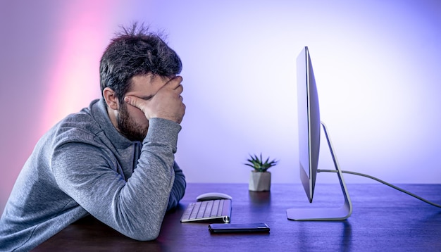 疲れた男がコンピューターの前に座って顔を手で覆っている