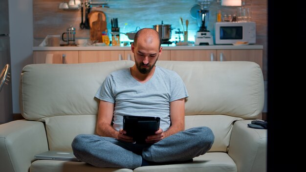 現代技術のワイヤレスを備えたタブレットコンピューターを使用してインターネット上でブラウジングしながらソファに一人で座っている疲れた男。キッチンで夜遅くにテレビジョンの前でリラックスしたパジャマを着た白人男性