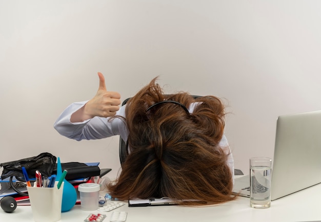 Tired middle-aged female doctor wearing medical robe and stethoscope sitting at desk with medical tools clipboard and laptop putting head on desk showing thumb up isolated