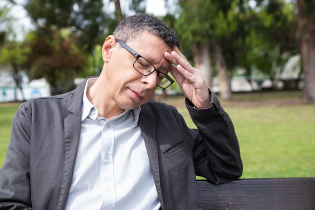 Tired middle-aged man touching head and sitting on bench in park