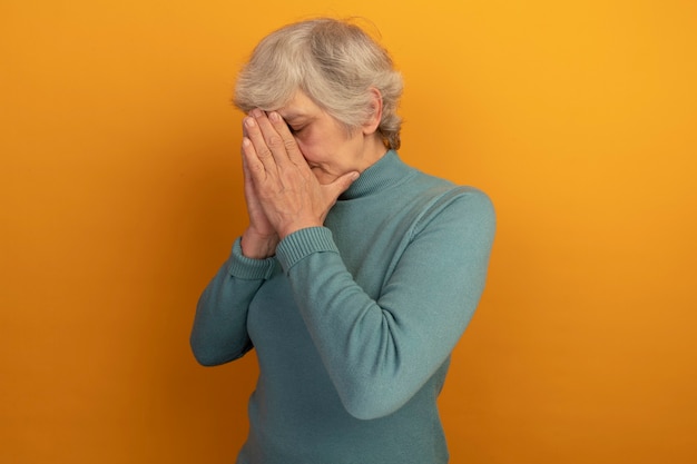 青いタートルネックのセーターを着て、コピースペースのあるオレンジ色の壁に隔離された目を閉じて顔に手を合わせて立っている疲れた老婆