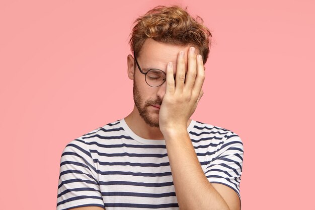 Tired overworked curly ordinary man keeps hand on face, closes eyes, feels sleepy