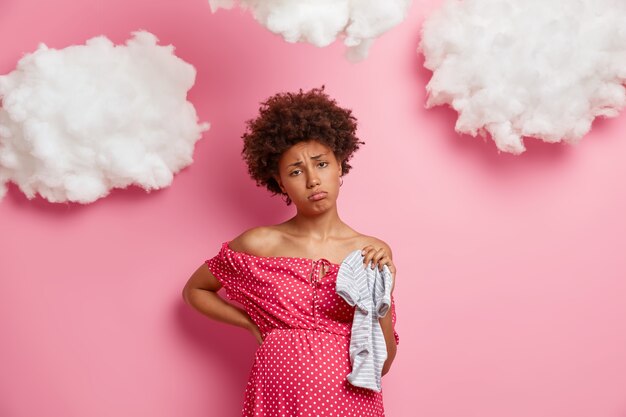 Tired pregnant woman massages her back, gets ready for newborn birth, holds clothes for baby, has displeased expression, fatigue after doing everyday homework, wears polka dot dress, isolated on pink