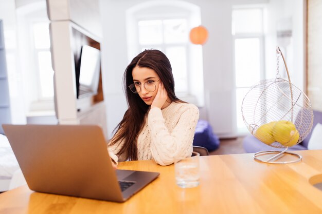 Tired pretty woman with lond black hair is working on her laptop and sitting at home at the wooden table