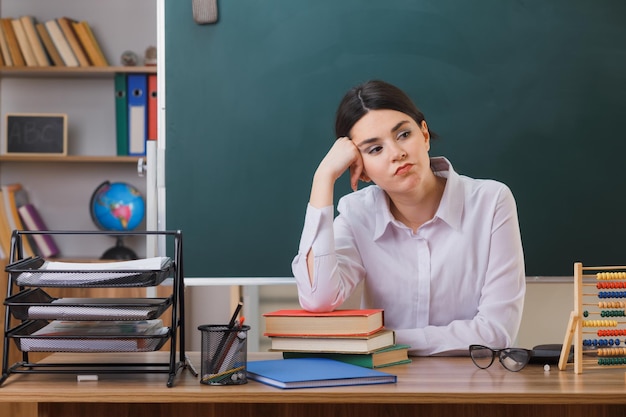 Free photo tired putting hand on cheek young female teacher sitting at desk with school tools in classroom