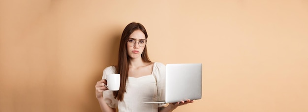 Free photo tired and reluctant girl hates working drinking morning coffee and using laptop frowning upset stand