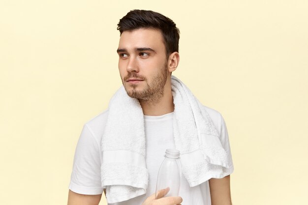 Tired serious young male having rest after cardio work out wearing white towel around his neck, drinking water from plastic bottle