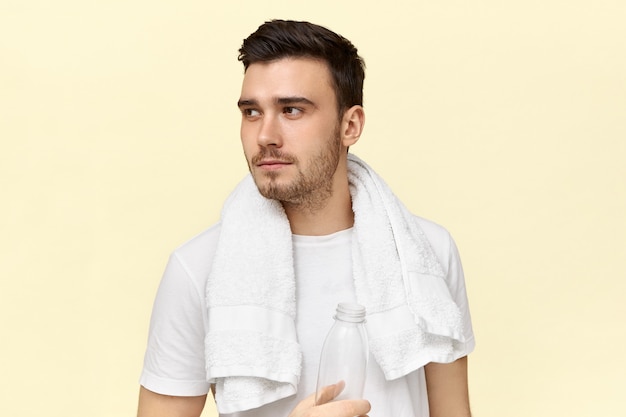 Tired serious young male having rest after cardio work out wearing white towel around his neck, drinking water from plastic bottle