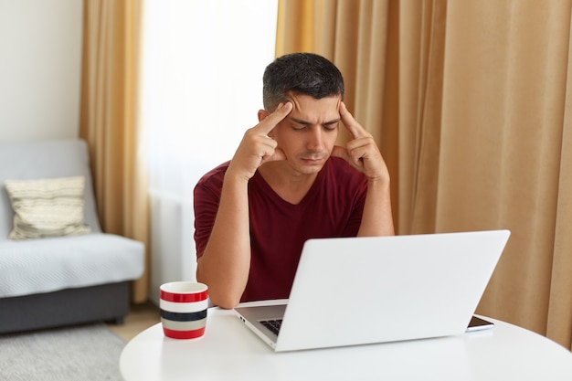 Усталый больной мужчина в повседневной одежде сидит за столом в гостиной, держит пальцы на висках, пытается расслабиться, страдает от головной боли после долгих часов работы за компьютером.