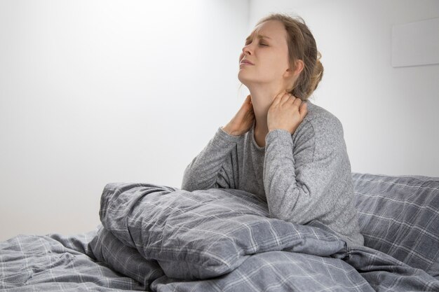 Tired sick woman on bed touching her neck, suffering from pain