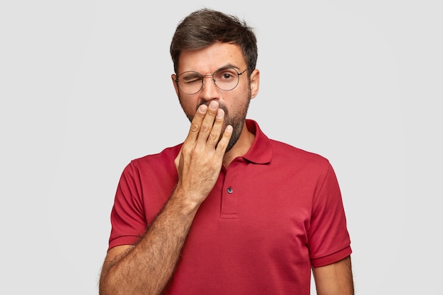 Tired sleepy bearded young male yawns, covers mouth with palm, wears spectacles and red t-shirt, stands against white wall, being fatigue after long work, isolated over white wall.