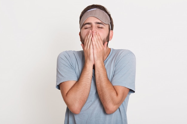 Tired sleepy bearded young man standing isolated over white wall in , yawning in morning, covering his mouth with hands