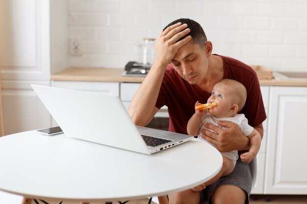 Tired sleepy handsome freelancer male wearing burgundy r shirt, posing in white kitchen, sitting in front of laptop with baby in hands, suffering terrible headache.