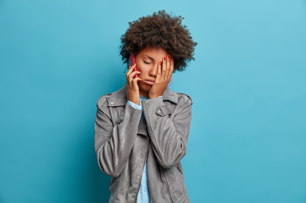 Free photo tired sleepy woman with afro hair makes face palm, feels tired and fatigue, has boring telephone conversation, calls friend, wears grey jacket, poses