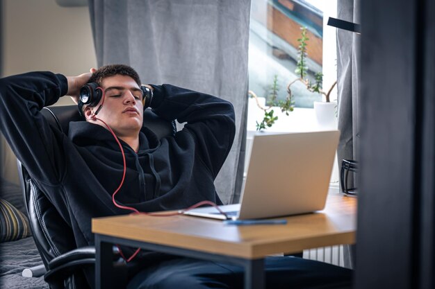 Усталый подросток в наушниках сидит перед ноутбуком в своей комнате