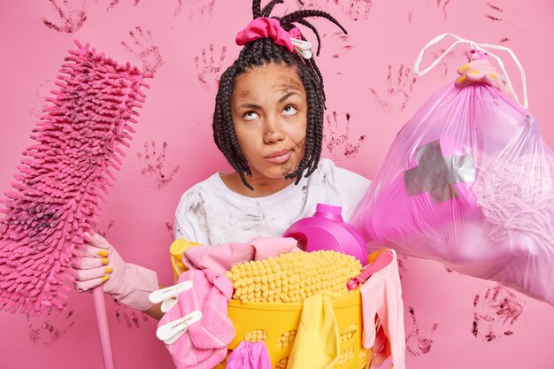 Tired thoughtful housewife poses with rubbish bag and mop stands dirty after doing housework cleans house uses cleaning products wears rubber protective gloves has dreadlocks isolated on pink wall