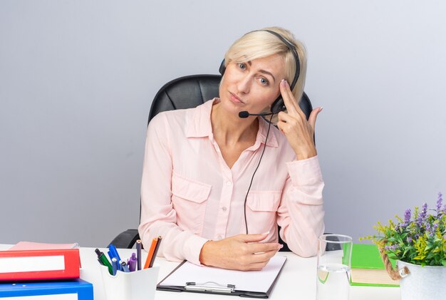 Tired tilting head young female call center operator wearing headset sitting at table with office tools showing suicide gesture isolated on white wall