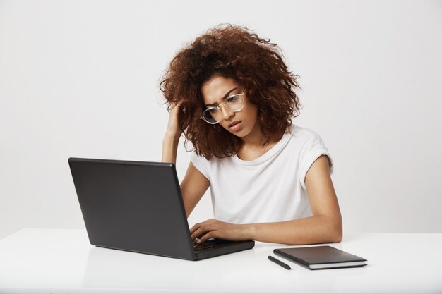 Tired and ucertain young african fashion design student thinking on her study working at laptop on business plan of future brand. Outstanding screenwriter in her twenties on white wall.