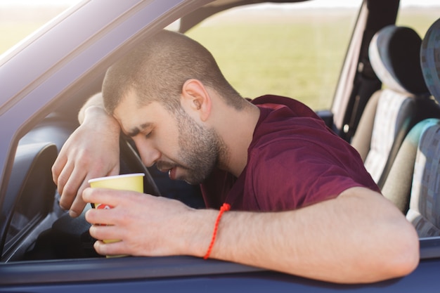 Il maschio con la barba lunga stanco si appoggia sulla ruota e tiene la tazza di caffè di carta