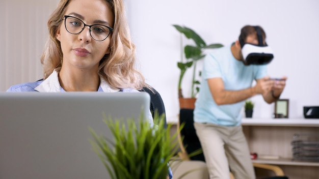 Donna freelance stanca e sconvolta che lavora al computer portatile in casa mentre suo marito la disturba giocando ai videogiochi con un visore vr in background.