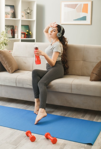 Tired with closed eyes putting hand on forehead young girl wearing headphones holding water bottle sitting on sofa in living room