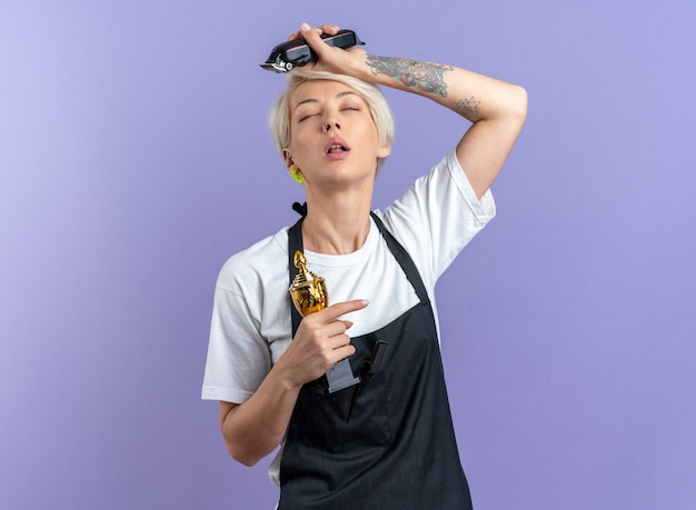 Tired with closed eyes young beautiful female barber in uniform holding winner cup with hair clippers isolated on blue background