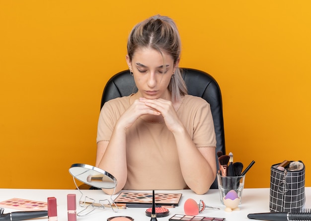 Tired with closed eyes young beautiful girl sits at table with makeup tools isolated on orange background