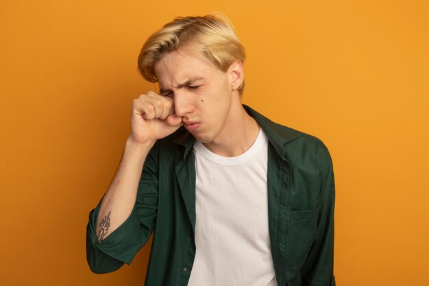 Tired with closed eyes young blonde guy wearing green t-shirt wiping eye with hand