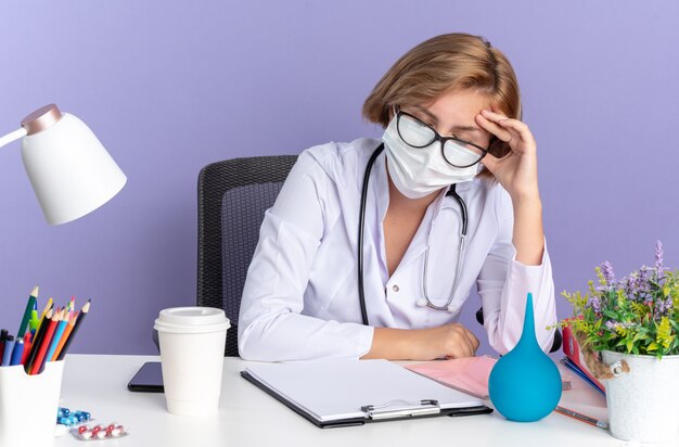 눈을 감고 피곤한 젊은 여성 의사는 청진기가 달린 의료 가운을 입고 의료 마스크가 달린 안경을 쓰고 파란색 배경에 격리된 의료 도구를 들고 탁자에 앉아 있다
