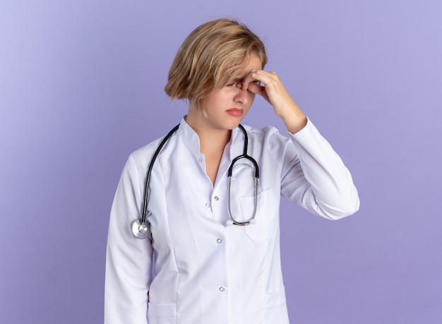 Tired with closed eyes young female doctor wearing medical robe with stethoscope grabbed nose isolated on blue background