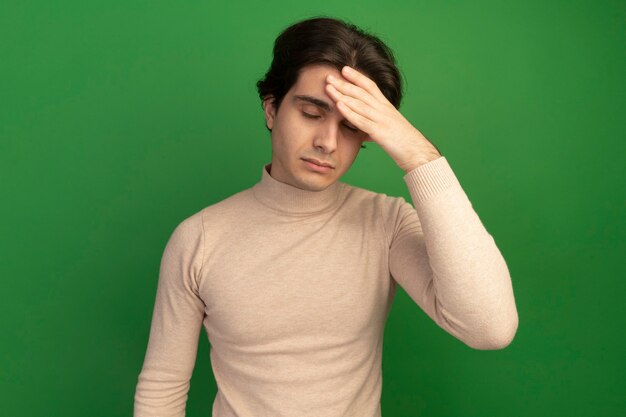 Tired with closed eyes young handsome guy putting hand on forehead isolated on green wall