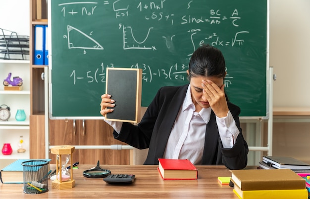 Tired with lowered head young female teacher sits at table with school tools holding mini blackboard in classroom