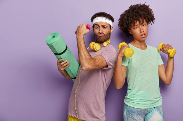Tired woman raises two dumbbells, works on biceps and determined unshaven man carries karemat for gymnastic training