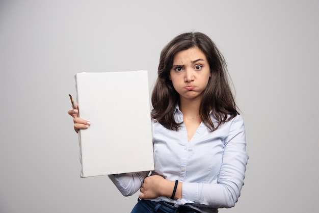 Tired woman showing empty canvas and paintbrush on gray wall. 