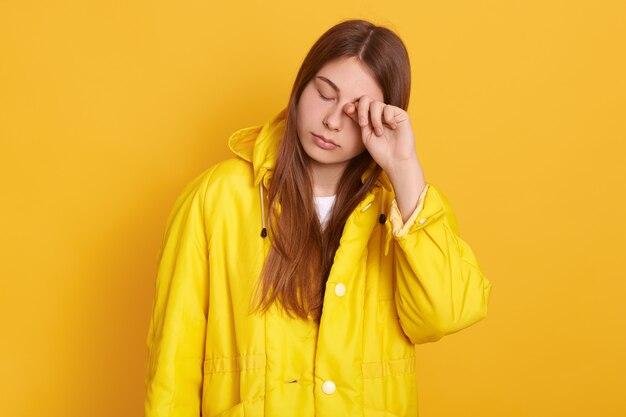 Tired woman wearing yellow jacket rubbing her eye, female with long beautiful hair posing with closed eyes, looks exhausted, standing against bright wall.