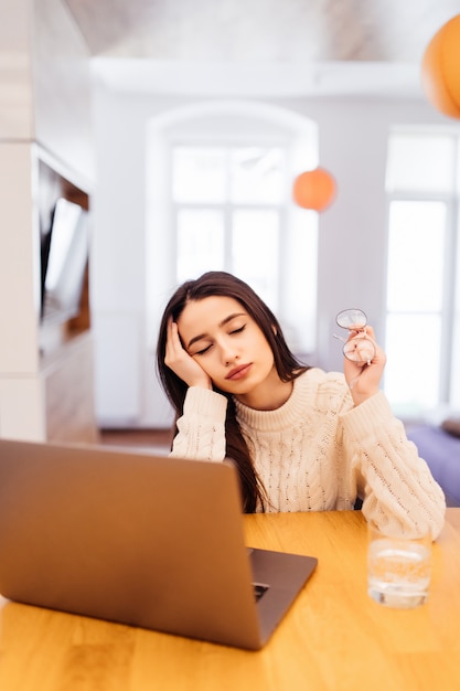 Tired worker with lond black hair is working on her laptop and sitting at home at the wooden table