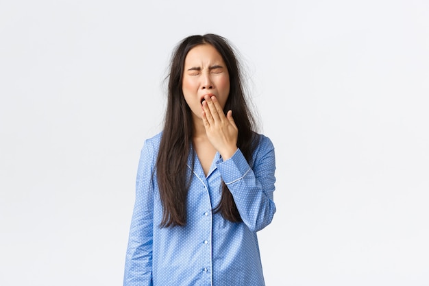 Tired yawning asian girl in blue pajamas, yawning sleepy with messy haircut after waking up, cover opened mouth with hand, standing in jammies after rough night, white background.