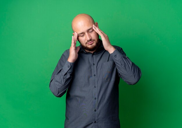 Tired young bald call center man putting hands on temples with closed eyes isolated on green wall