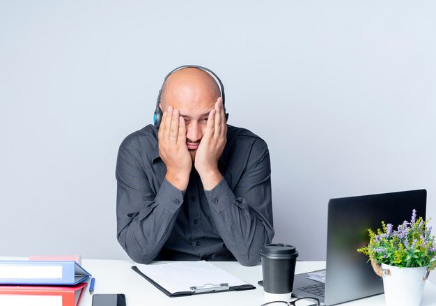 Tired young bald call center man wearing headset sitting at desk with work tools putting hands on face isolated on white wall