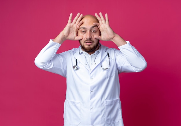 Tired young bald male doctor wearing medical robe and stethoscope opening eyes with hand isolated on pink