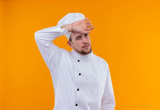 A tired young bearded chef man in white uniform holding hand on head while looking on an orange wall