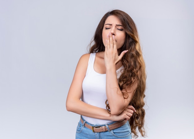 Tired young beautiful girl putting hand on mouth and yawning with closed eyes on isolated white wall with copy space