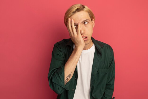 Tired young blonde guy wearing green t-shirt covered face with hand