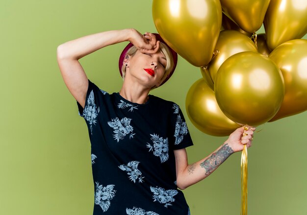 Tired young blonde party girl wearing party hat holding balloons touching forehead with closed eyes isolated on olive green background with copy space