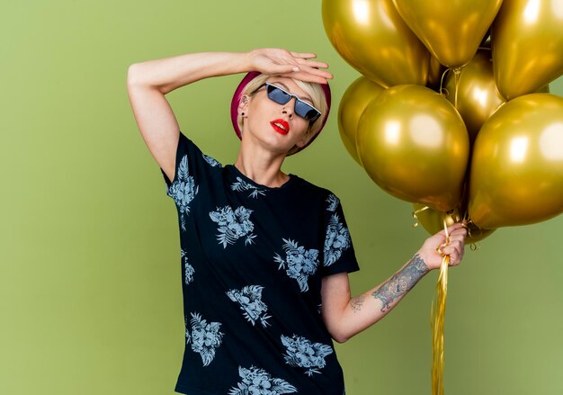 Tired young blonde party woman wearing party hat and sunglasses holding balloons putting hand on forehead isolated on olive green wall with copy space