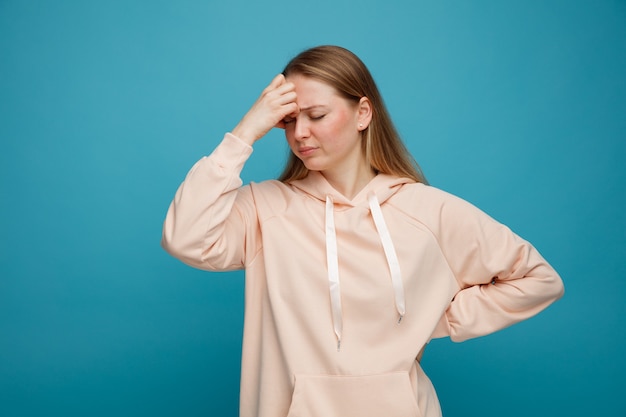 Tired young blonde woman keeping hand on forehead and on waist with closed eyes 