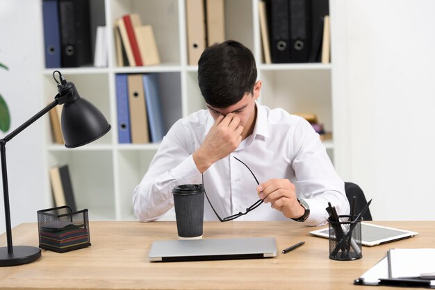 Tired young businessman holding eyeglasses in hand with takeaway coffee cup and laptop on desk