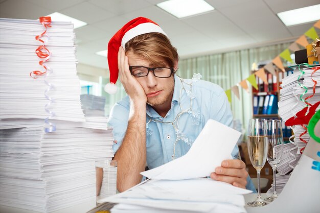 Tired young businessman sitting at workplace among papers on christmas day.